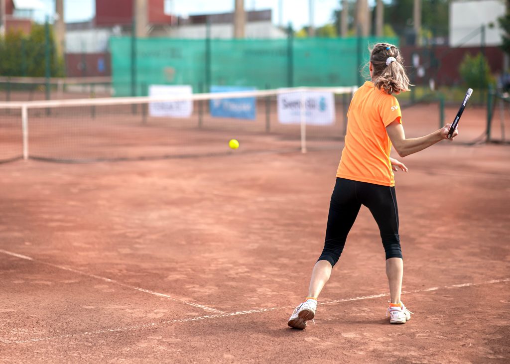 Woman playing tennis on clay court wearing orange sportswear, back view.